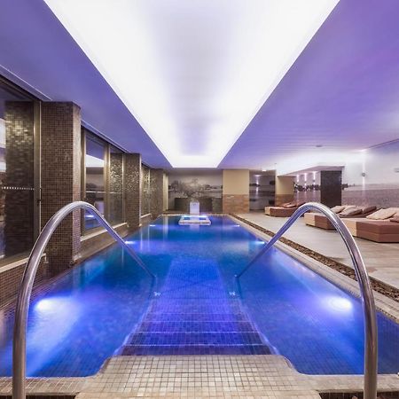 Ap Lago Montargil - Conference & Spa Екстер'єр фото The photo shows a modern indoor swimming pool area. The pool features a sleek design with steps leading into the water and a gentle curve at the sides. The water is illuminated in a blue hue, enhancing the relaxing atmosphere. Surrounding the pool ar