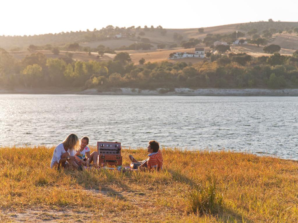 Ap Lago Montargil - Conference & Spa Екстер'єр фото The photo depicts a serene lakeside scene during sunset. Three people are gathered on a grassy area near the water, enjoying a picnic. They are seated around a picnic basket, with the shimmering lake in the background and hills in the distance. The w