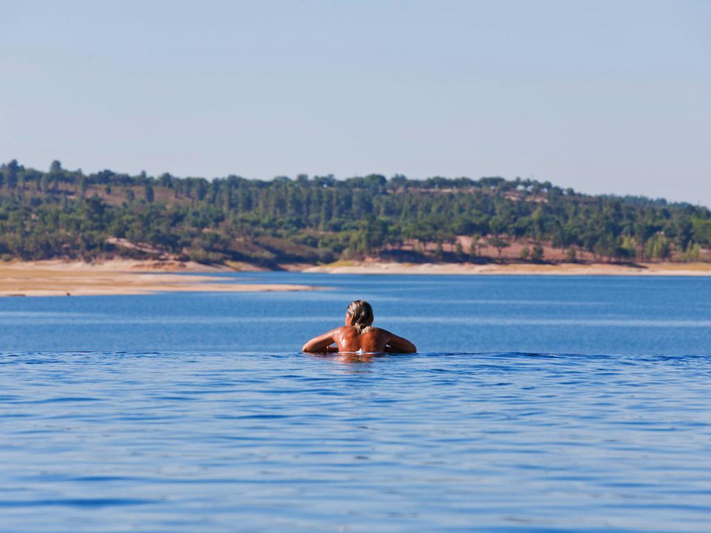Ap Lago Montargil - Conference & Spa Екстер'єр фото The photo shows a person swimming in a calm body of water, likely a lake or a river. The swimmer is seen from behind and appears to be enjoying the water on a sunny day. In the background, there's a scenic landscape featuring green trees and a sandy 