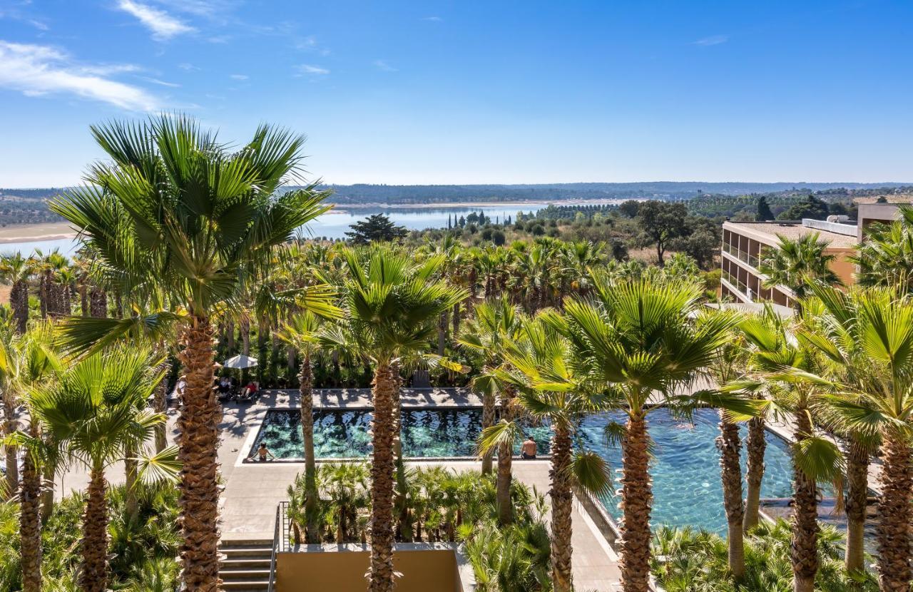 Ap Lago Montargil - Conference & Spa Екстер'єр фото The photo depicts a picturesque resort landscape with a clear blue sky. In the foreground, there are tall palm trees surrounding a swimming pool area. The swimming pool has a modern design and is surrounded by lush greenery. In the background, you ca
