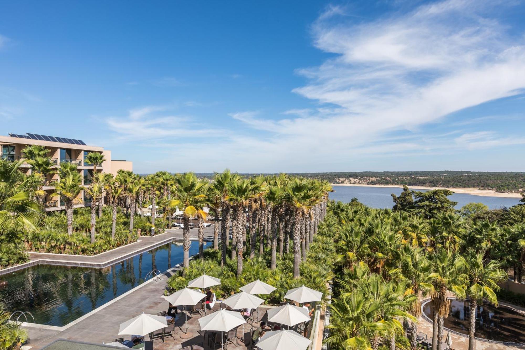 Ap Lago Montargil - Conference & Spa Екстер'єр фото The photo shows a scenic view of a resort or hotel area. In the foreground, there is a pool surrounded by palm trees and lounge chairs with umbrellas, suggesting a relaxing atmosphere. Beyond the pool, there is a body of water, likely a river or coas