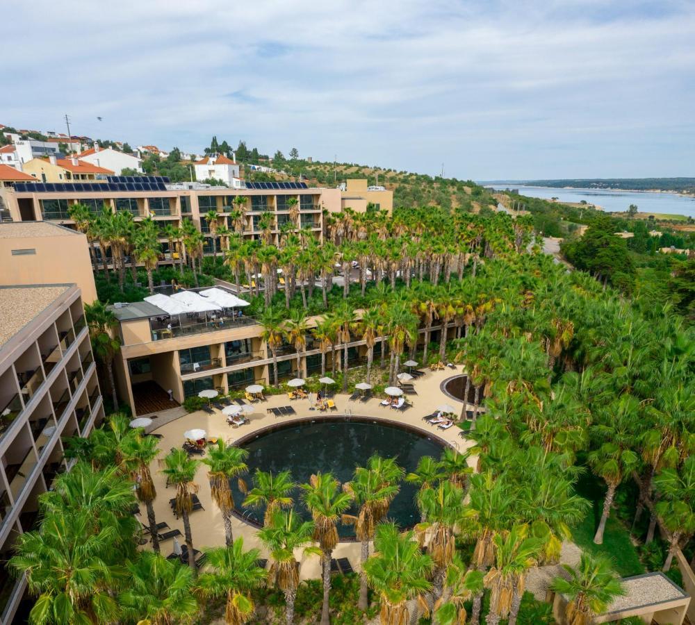 Ap Lago Montargil - Conference & Spa Екстер'єр фото The photo shows a modern hotel complex set amidst lush greenery, featuring numerous palm trees. In the foreground, there is a circular swimming pool surrounded by lounge chairs. The hotel itself has multiple levels with balconies overlooking the pool