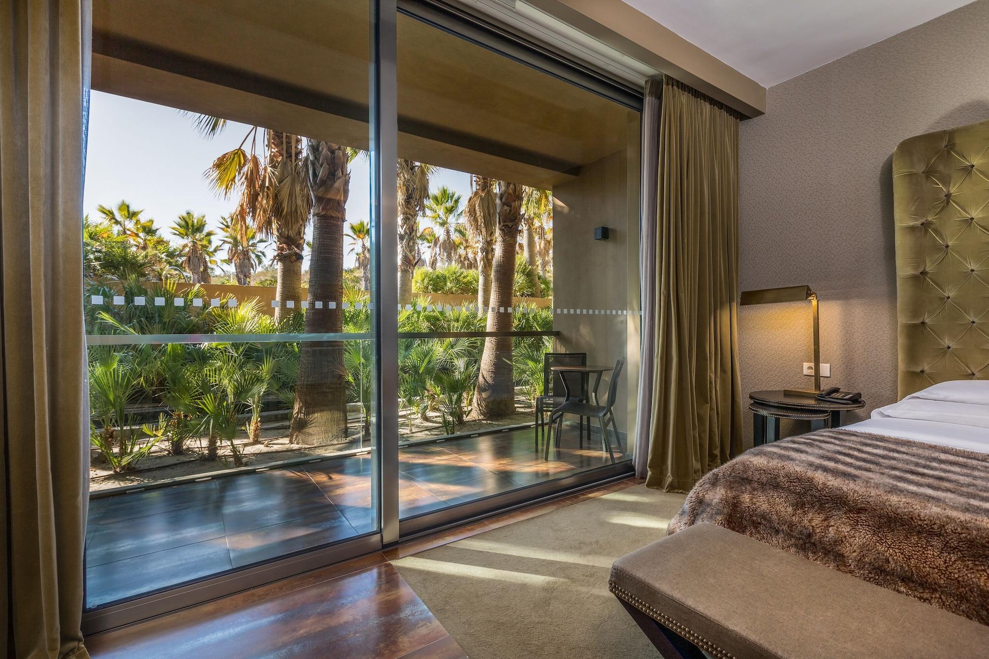 Ap Lago Montargil - Conference & Spa Екстер'єр фото The photo shows a bedroom with large glass doors that lead out to a balcony or terrace. The view outside features palm trees and lush greenery, indicating a tropical or warm climate. Inside the room, there is a bed, a bedside table with a lamp, and a