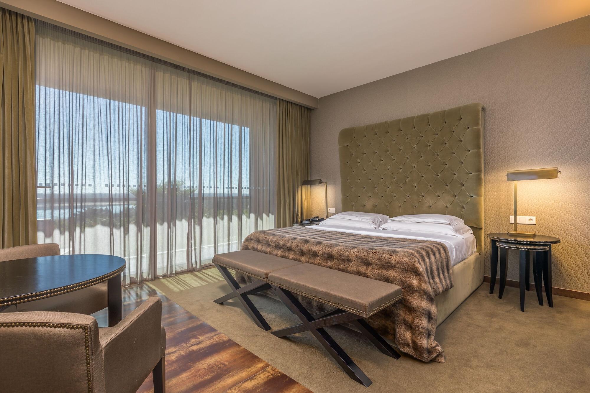 Ap Lago Montargil - Conference & Spa Екстер'єр фото The photo shows a modern hotel room featuring a large bed with a tufted headboard and a cozy blanket. There are curtains draped over large windows that allow natural light to flow in, providing a view of the outside. A small seating area includes two