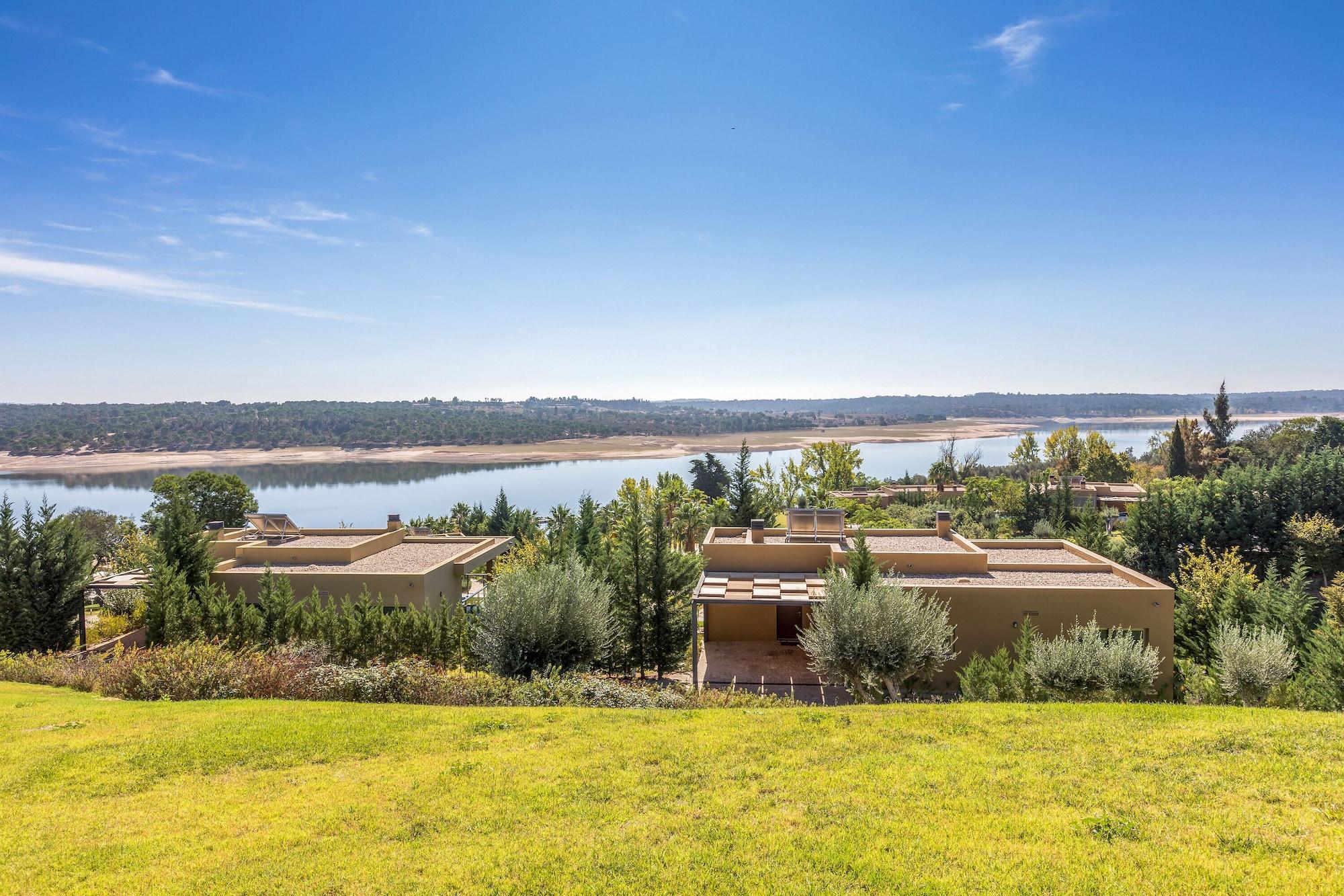 Ap Lago Montargil - Conference & Spa Екстер'єр фото The photo depicts a scenic landscape featuring a calm body of water, possibly a lake or river, surrounded by lush greenery and trees. In the foreground, there are several modern buildings with flat roofs, set against a vibrant blue sky. The overall s