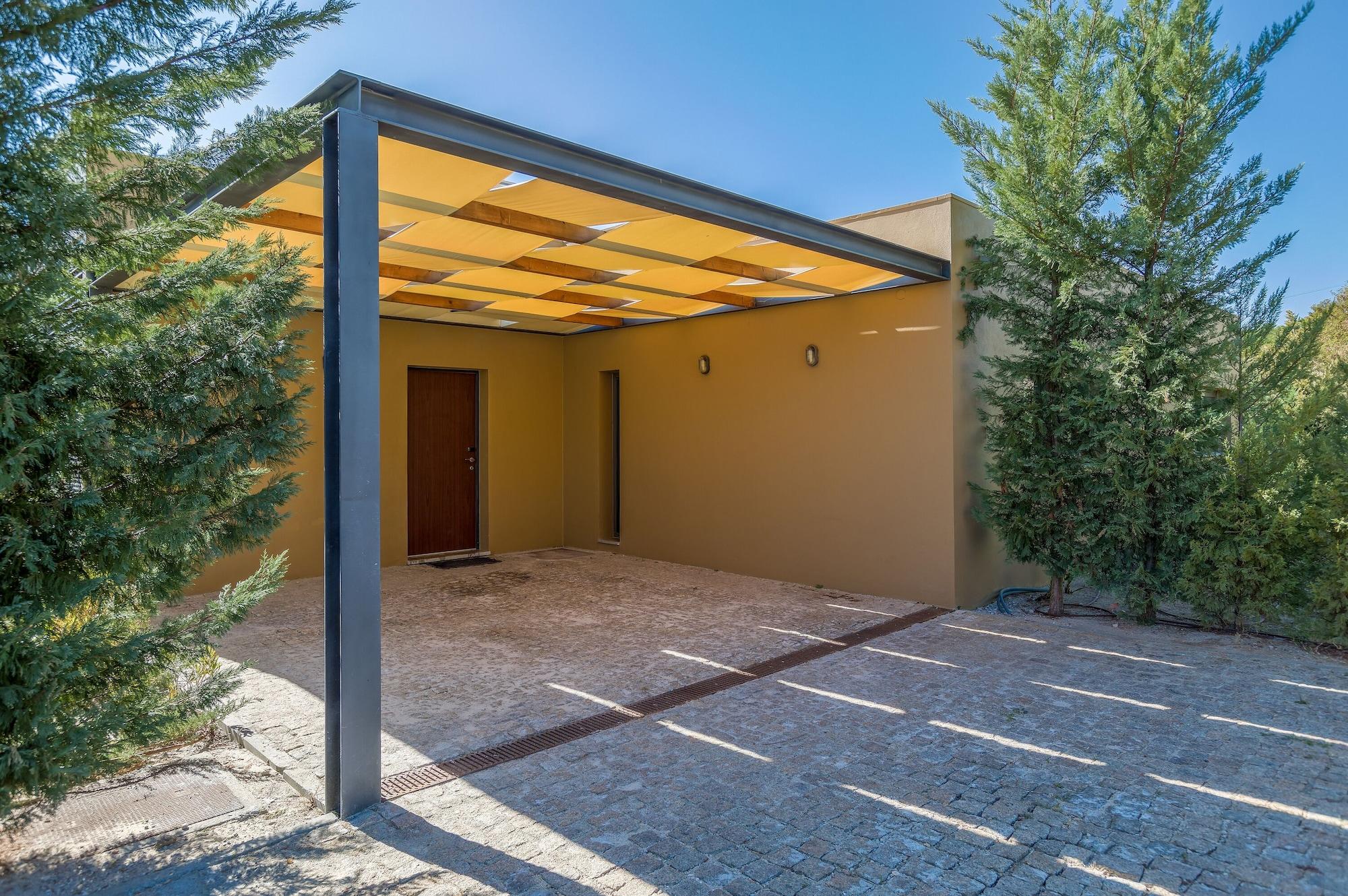 Ap Lago Montargil - Conference & Spa Екстер'єр фото The photo shows an exterior view of a modern structure with a carport. The carport has a sleek, metallic frame and a yellowish canopy above it. In front of the structure, there is a paved area made of stone or tiles. The main entrance features a wood