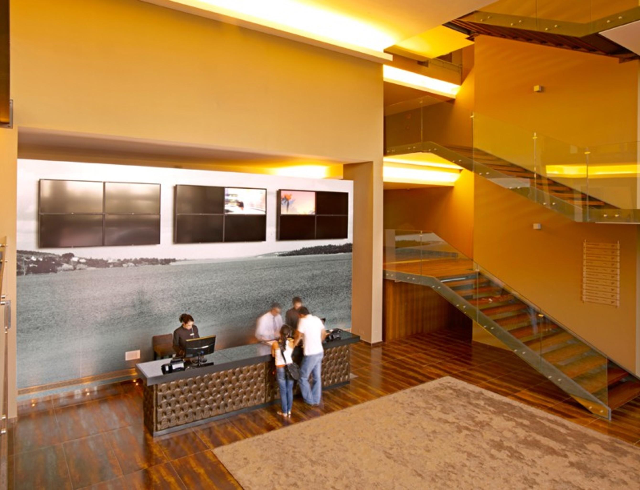 Ap Lago Montargil - Conference & Spa Екстер'єр фото The photo depicts a modern lobby area with a reception desk. The design features a combination of warm colors and wood flooring. In the background, there is a large wall-mounted screen or display, showcasing several monitors. The wall behind the desk