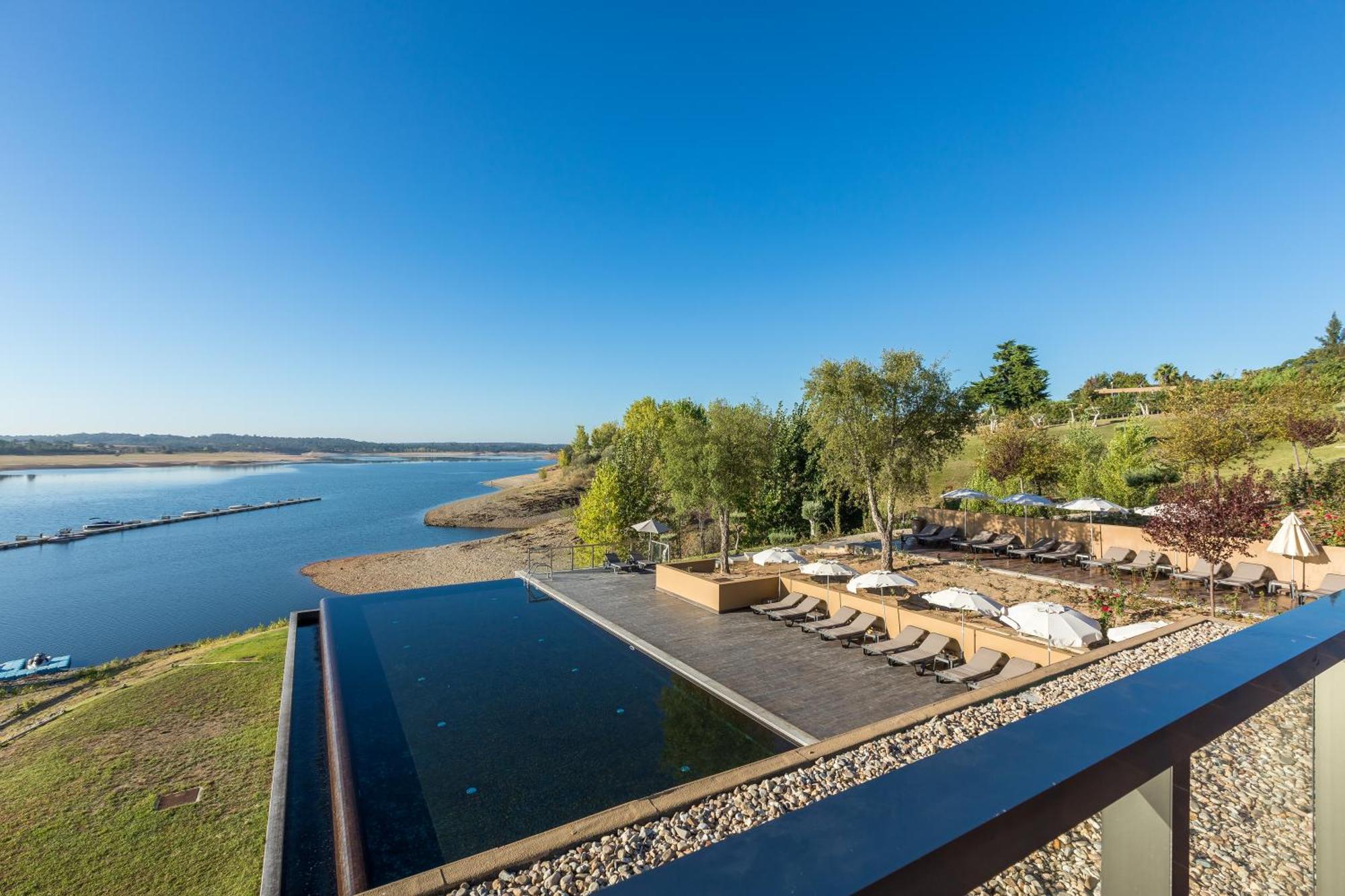 Ap Lago Montargil - Conference & Spa Екстер'єр фото The photograph depicts a scenic view of a tranquil water body, likely a lake or river, bordered by lush greenery. In the foreground, there is an infinity pool that blends seamlessly with the water beyond. Comfortable lounge chairs are arranged on a w
